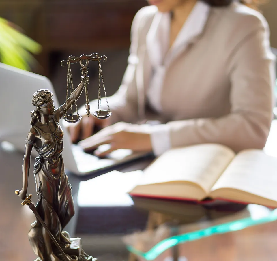 Bronze statue of Lady Justice holding scales and a sword, with a woman working at a desk in the blurred background.